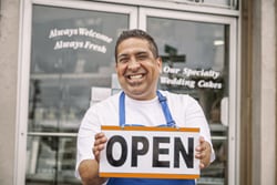 Smiling local small business owner stands in front of store with open sign after placing digital ad