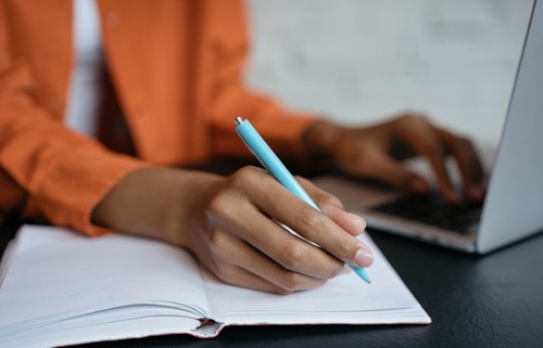 Close-up shot of hand holding pen and writing in notebook while using a laptop for content creation