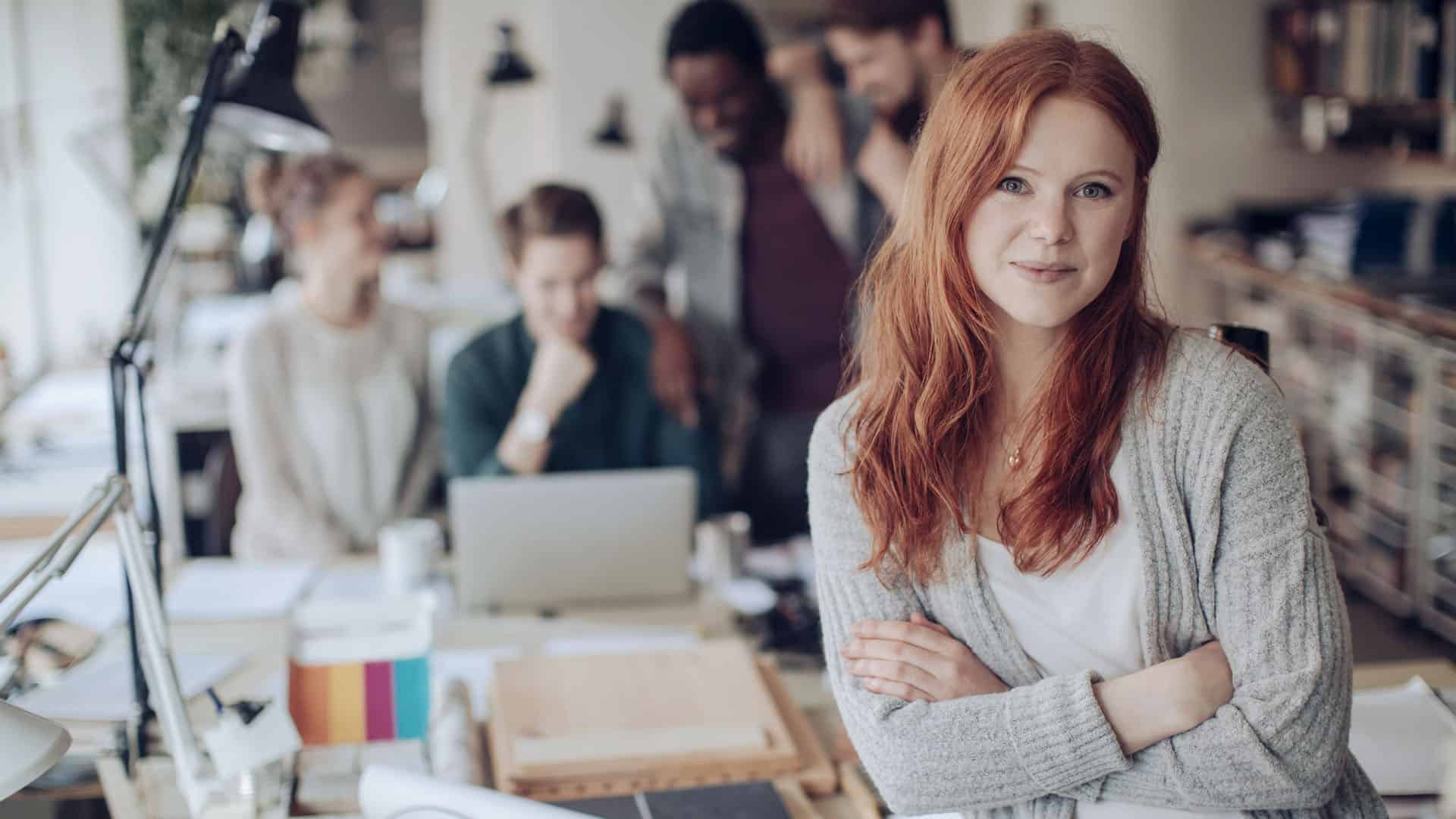 Close up young woman looking at camera with her digital marketing agency colleagues in background