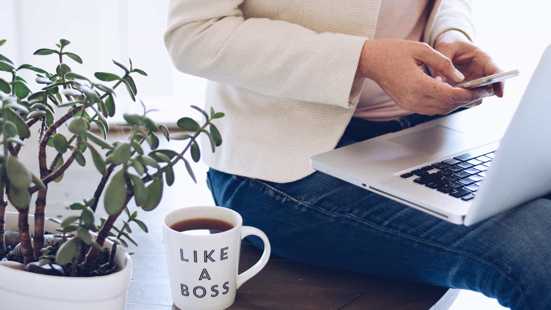 A small business owner texting while sitting on a desk with a coffee cup that says ‘Like a Boss’