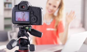Young woman smiling and waving at camera recoding a video for her YouTube video marketing channel