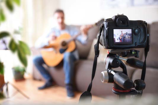 Camera recording video blogger doing a review of a guitar in the living room for ecommerce website