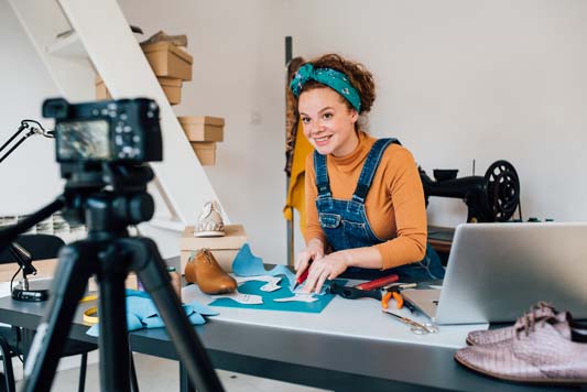 Young woman cutting leather while filming educational video about making shoes in her workshop