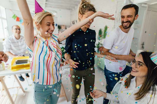 Happy coworkers with party hats throwing confetti in office to celebrate customer appreciation week