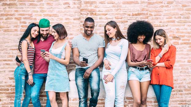 Happy multiethnic friends leaning against brick wall using smartphones to check email communication