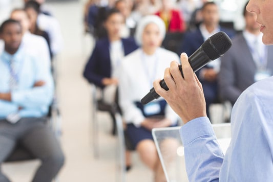 Side view of female speaker with microphone in hand facing a large group of seated business people