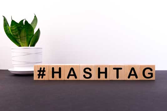The word hashtag spelled out on a desk using scrabble letters with a plant in the background