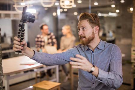 Male entrepreneur making a selfie video blog by holding a digital camera in front of face at office