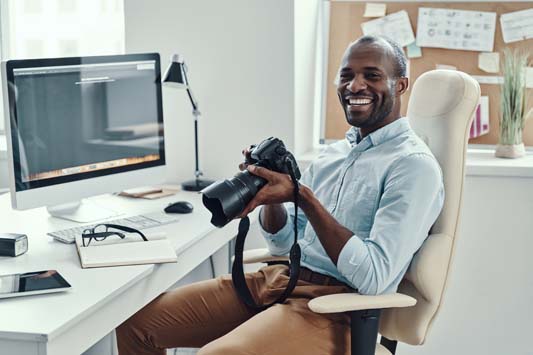 Smiling male business owner sitting at desk holding digital camera to review product photography