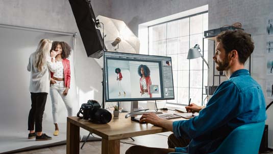 Make-up artist applies makeup on model as photo editor works on desktop computer retouching photos