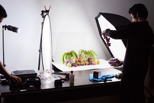 Food product photography setup on white backdrop with lighting reflector while assistant adds fruit