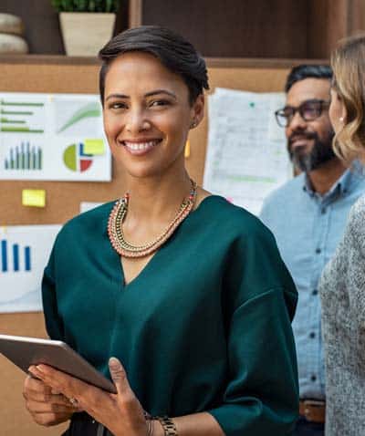 Outsourced expert with digital tablet smiling at camera while owners in background review progress