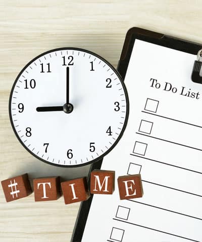 Overhead of clock on desk with blocks spelling out time on top of a clipboard with a to do list